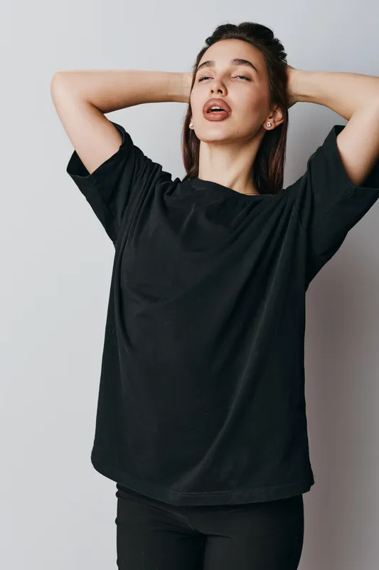 Young woman in black t shirt posing with hands behind head and eyes closed on gray background. Casual style, relaxed expression and natural beauty in studio portrait.