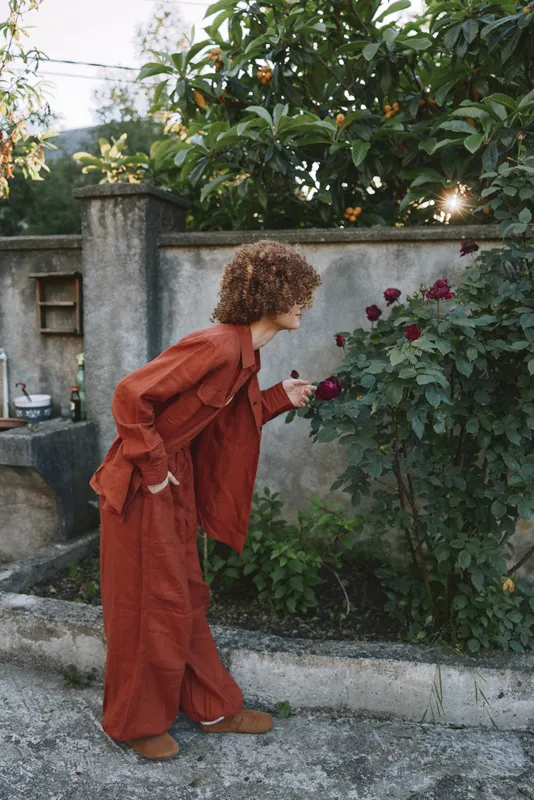 A woman in an orange rust jumpsuit leans toward a rose bush in a quiet courtyard, gently smelling blossoms and smiling with calm focus
