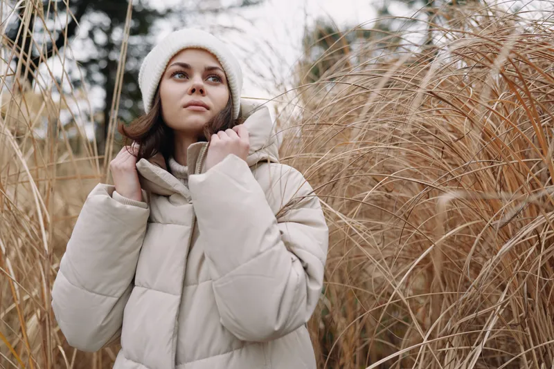 Winter scene with a woman in a puffer coat and knit hat among tall grasses, a calm outdoor portrait conveying thoughtful mood and natural seasonal beauty.