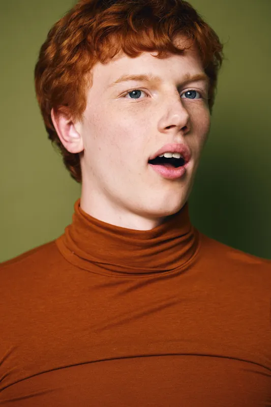 Young redhead man in brown turtleneck with freckles and open mouth posing against green background looking surprised or curious in studio portrait closeup.