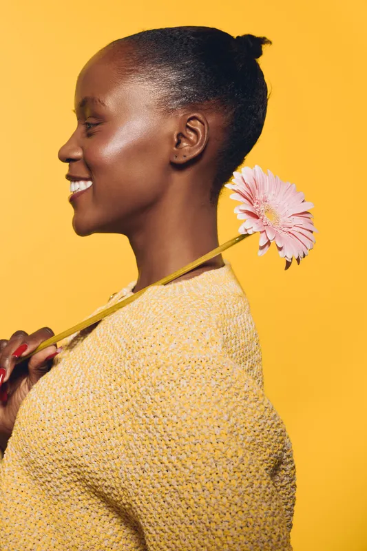 Profile of a smiling black African woman with dark skin holding a pink flower on her shoulder. She wears a yellow knit sweater against a bright yellow background. Concept of happiness and beauty.