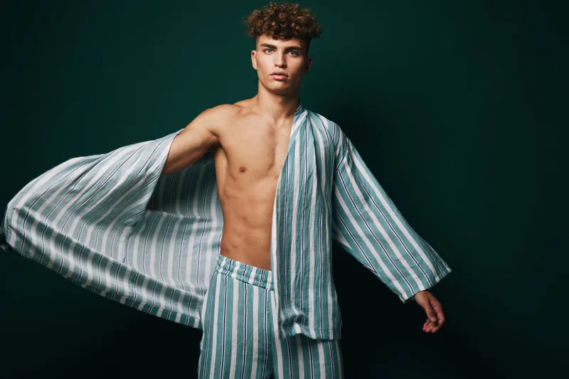 Young man with curly hair wearing striped pajamas and robe posing against dark background for fashion, style and casual homewear concept with confident expression.