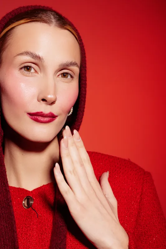 Close-up portrait of a woman with clear skin and red lips wearing a red sweater and hood against a red background. Concept of beauty, fashion, and skincare.