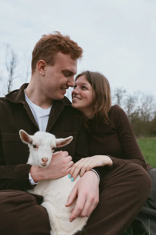 Lifestyle shot of a couple outdoors with a friendly goat kid, smiling and affectionate, sitting in a field, enjoying rural life, warm bond and relaxed moment, natural light, candid vibe, simple
