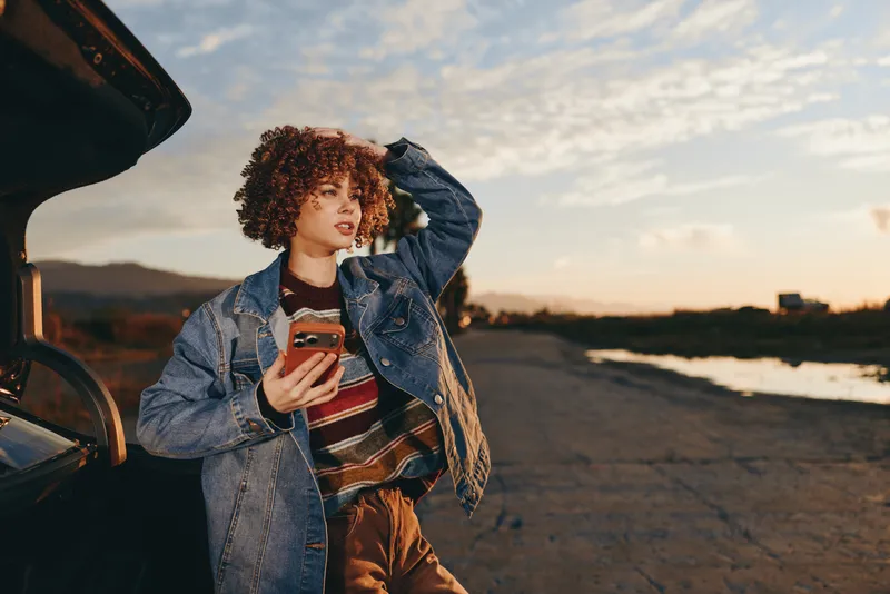 Woman outdoors at sunset, lifestyle moment by an open car trunk, curly hair, holding phone, denim jacket and casual smile, warm evening light, road and distant hills, soft clouds, golden sky