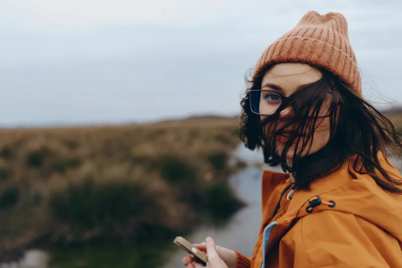 Woman lifestyle outdoors in autumn wind, wearing hat, glasses, orange coat and scarf, holding a smartphone, looking sideways toward a blurred hillside, casual travel moment