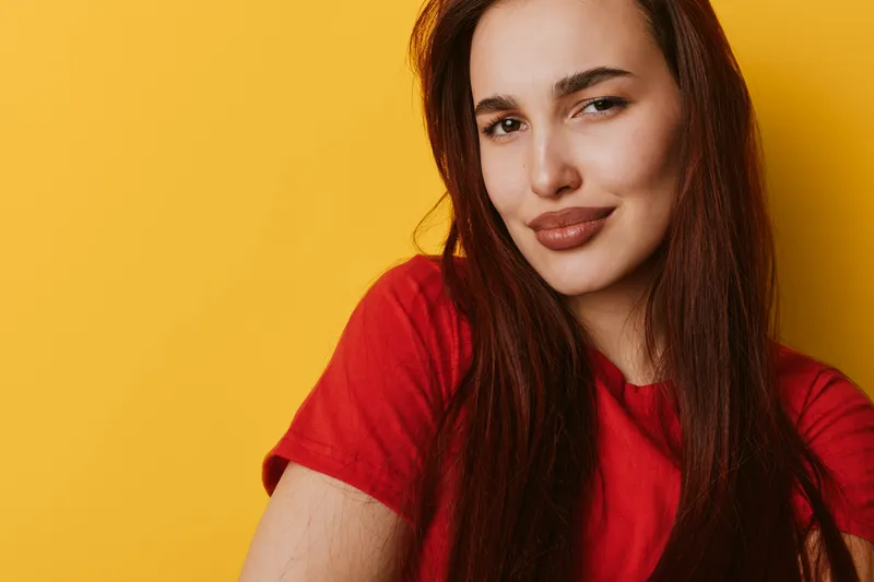 Woman with long brown hair wearing red shirt posing smiling confidently against yellow background in studio. Portrait of female model with natural makeup and casual style in bright setting.