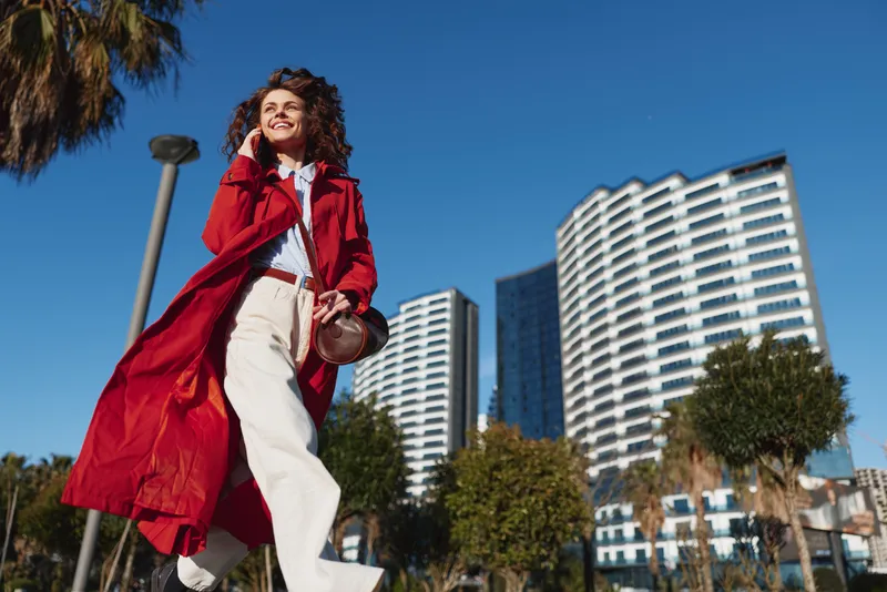 Stylish woman in a red coat walks through a sunny urban plaza, smiling confidently, as modern high rise buildings rise behind her.
