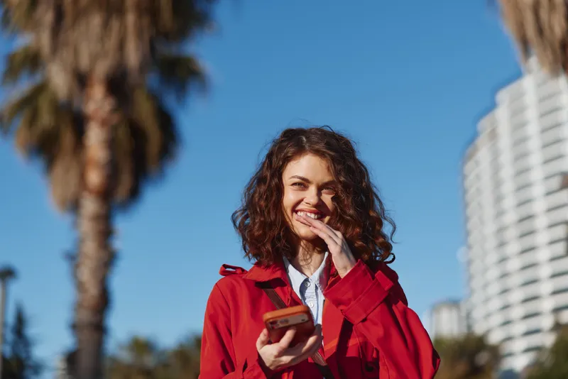 Young woman with curly hair wearing a red jacket smiles while holding a smartphone outdoors on a sunny day with palm trees in the background.