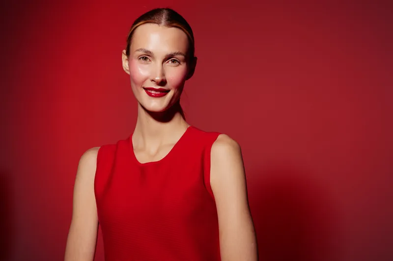 Smiling woman in red sleeveless dress poses against red background. Studio portrait with soft lighting highlights natural beauty and confident expression. Concept of fashion and elegance.