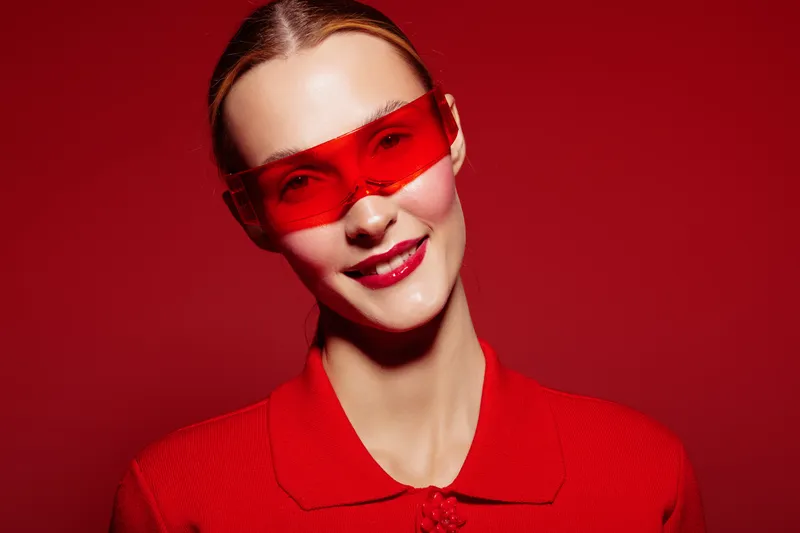 Smiling woman with red sunglasses and red outfit poses against red background. Fashion portrait with vibrant colors and confident expression. Concept of style and modern accessories.