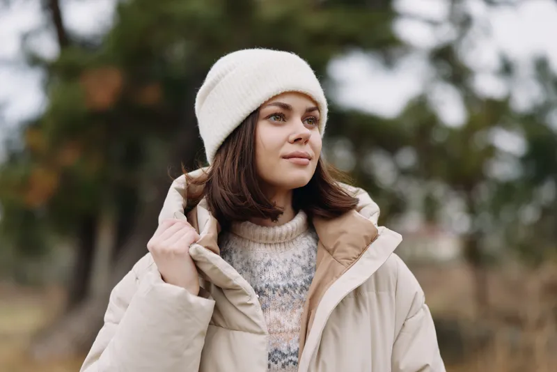 Woman portrait in outdoor setting wearing a beige coat and knit hat, calm expression, soft light, autumn backdrop, cozy winter fashion shot for lifestyle and apparel