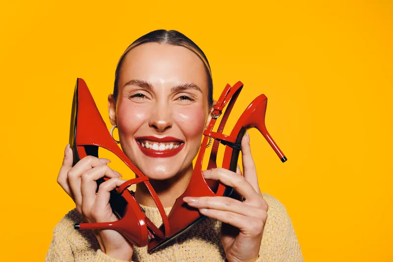 Smiling woman holds red high heel shoes near face on bright yellow background. Concept of fashion, happiness, style, and vibrant color contrast in studio portrait.