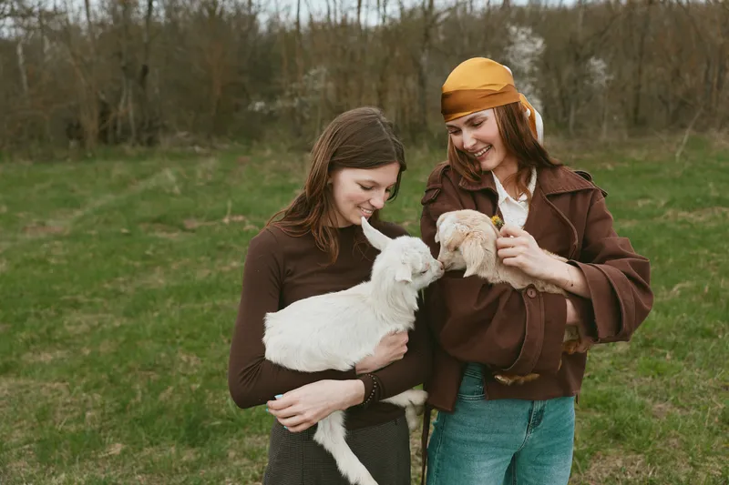 Lifestyle scene with two women outdoors holding a kid goat, sharing a gentle moment. Natural light, grassy field, casual wear, connection with animals, promoting wellness and friendship.