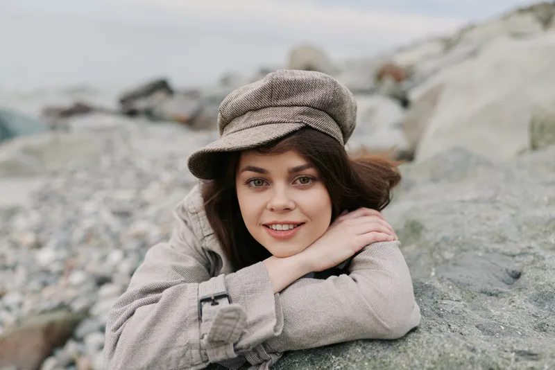 Smiling young woman in checkered coat and cap lies on rocky beach, resting head on crossed arms. Casual autumn fashion and natural beauty concept with soft natural light and serene mood.