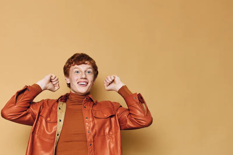 Young man in orange leather jacket and turtleneck sweater smiling with raised fists on beige background. Casual style, happy expression and confident positive gesture portrait.