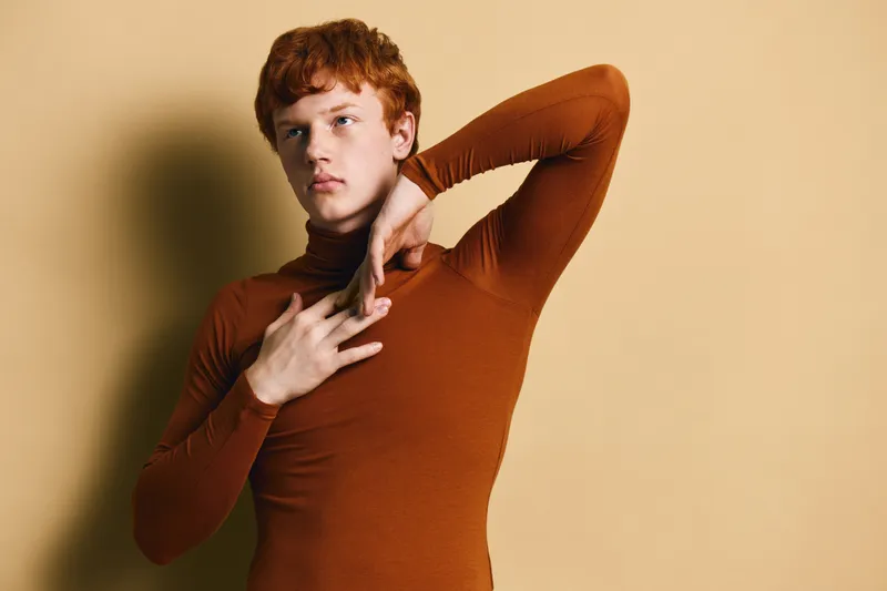 Young man with red hair wearing brown turtleneck posing with raised arm and hand on chest against beige background in studio portrait with soft lighting.
