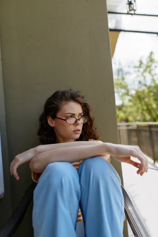 Young woman with glasses sits against a light concrete wall on a sunlit balcony, knees drawn up, exploring quiet, reflective mood in a relaxed home lifestyle moment