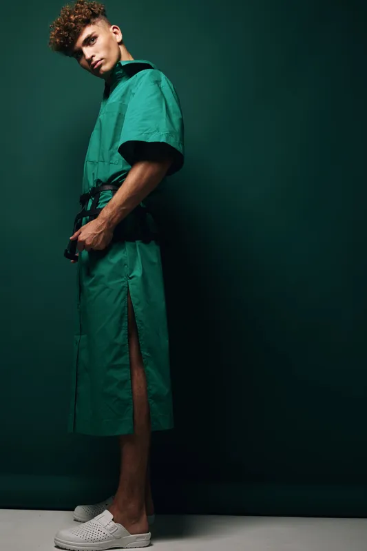 Young man in green medical scrubs standing against dark background with hands in pockets and white shoes, posing confidently in studio lighting.