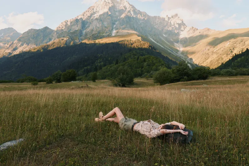 Cheerful woman outdoors lying in a grassy field with distant mountains, sunlit moment, relaxed posture, gentle breeze, natural colors, lush scenery