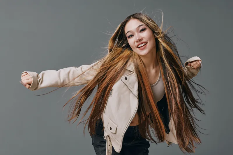 Young woman with long hair smiling and leaning forward with open arms wearing beige jacket and casual clothes on gray background in studio. Happiness and youth expression.