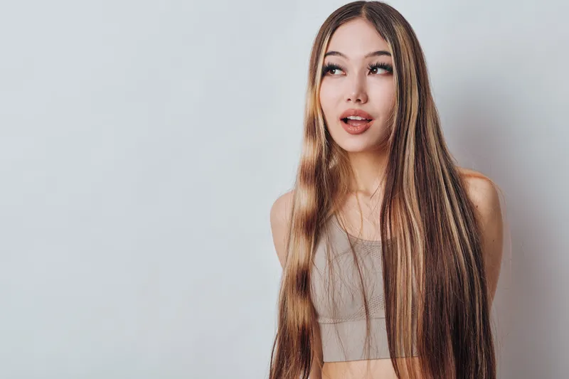 Young woman with long braided hair wearing a sleeveless top standing against a light background looking sideways with a thoughtful expression. Beauty and fashion concept.