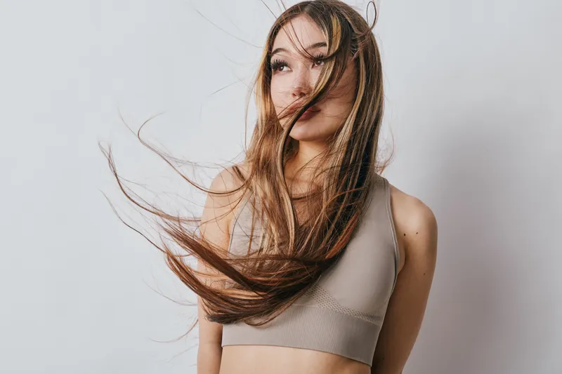 Young woman with long flowing hair wearing casual beige tank top looks away while hair moves in soft wind against plain white background in studio setting for beauty or fashion.