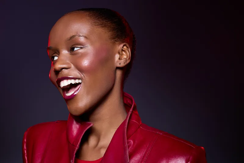 Smiling African woman with dark skin wearing red leather jacket looks sideways with open mouth. Studio portrait on dark background. Concept of fashion, joy, and beauty with vibrant colors.