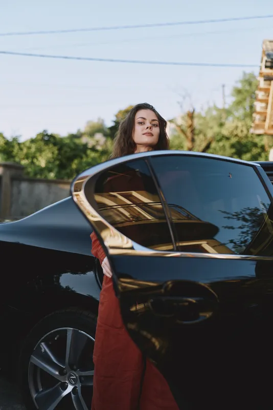 Curious woman stands beside a sleek black car in an outdoor setting, embodying a confident lifestyle moment, stylish fashion, and everyday chic under bright daylight