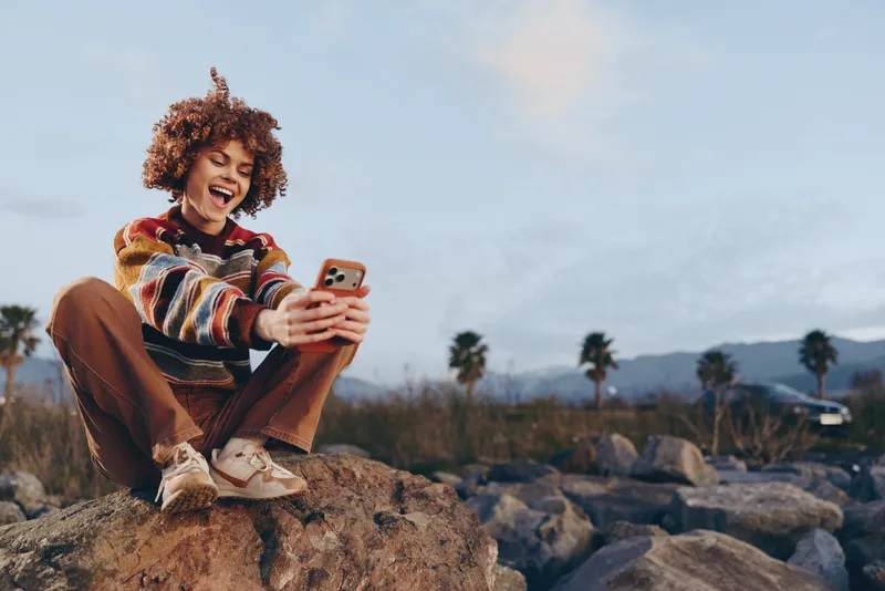 Woman outdoors lifestyle smiling curly hair, seated on large rocks, holding smartphone, laughing in bright daylight, casual travel vibe, palm trees and desert landscape, natural colors, relaxed