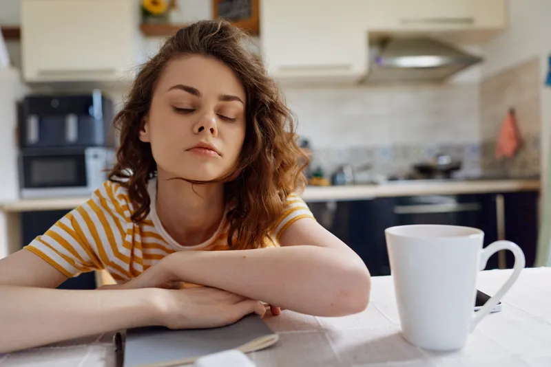 Calm woman resting on a table in a cozy kitchen, striped shirt, laptop nearby, mug on the surface, soft light creates a warm, relaxed home moment showcasing simple self care