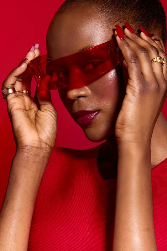 Close-up of a stylish black African woman with dark skin wearing red sunglasses and matching red top. Concept of fashion, beauty, confidence, and modern style on red background.