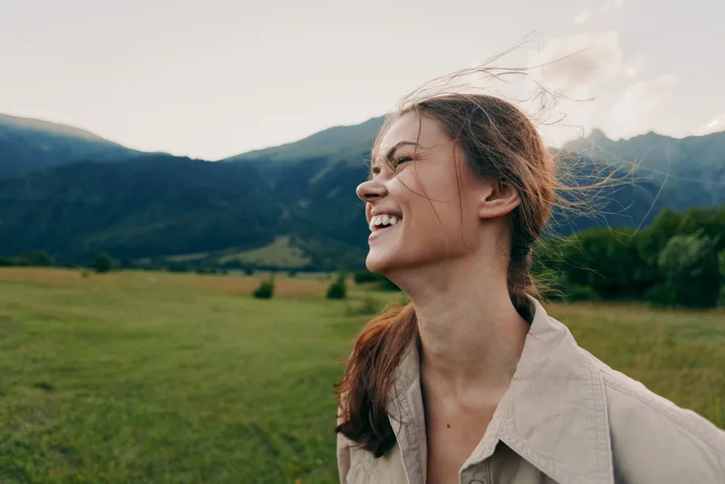 Smiling woman outdoors in a field with distant mountains. Natural light creates a warm, carefree mood and highlights lifestyle, outdoors, nature, beauty, happiness.