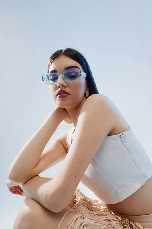 Young woman with long dark hair wearing stylish blue sunglasses and white crop top poses confidently against a light background. Concept of woman beauty fashion and style.