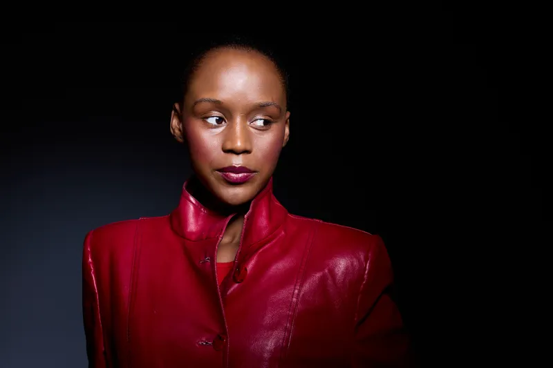 Portrait of an elegant black woman with dark skin wearing a shiny red jacket on black background. Studio lighting highlights her confident expression and smooth skin texture.