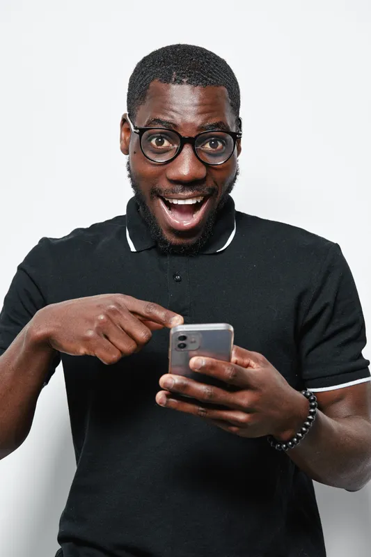 Black man with dark skin and glasses wearing black polo shirt smiling excitedly while pointing at smartphone screen isolated on white background. Technology, communication and happiness concept.