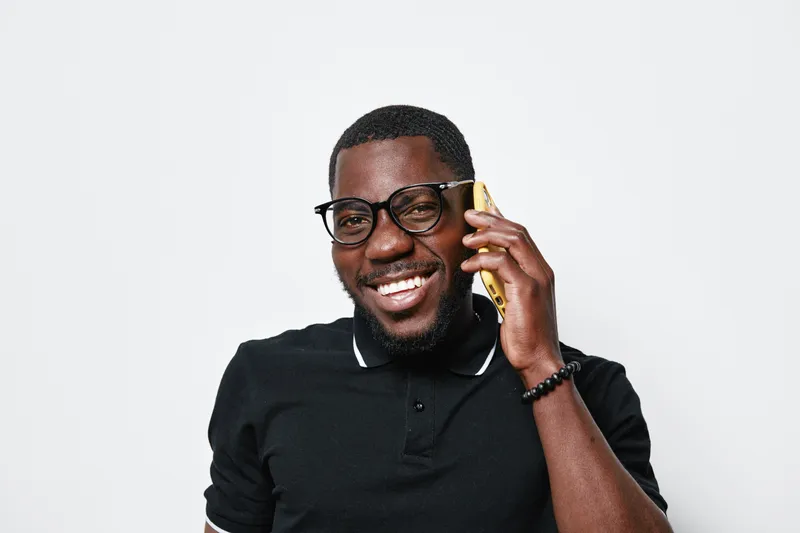 African American man with dark skin wearing glasses and black polo shirt smiling while talking on smartphone against white background. Positive communication and technology concept.
