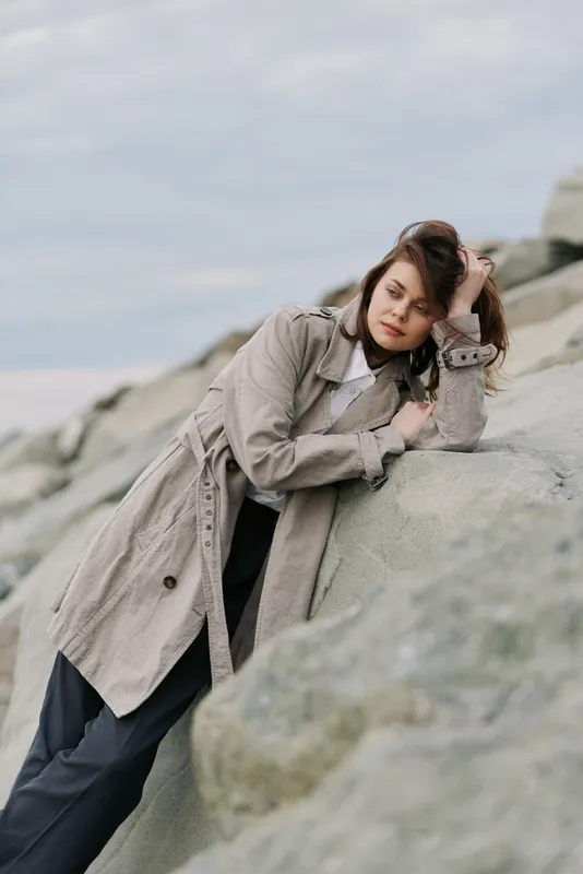 Young woman in beige trench coat leaning on rocks outdoors, looking thoughtful. Overcast sky and natural stone background create calm, reflective mood and autumn fashion concept.