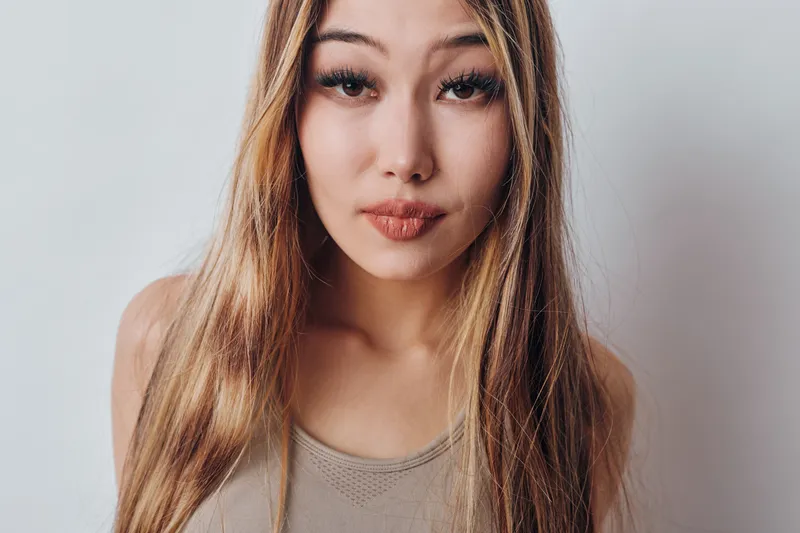 Young woman with long hair and natural makeup looking at camera on white background. Portrait shot with expression and casual tank top in studio setting with soft lighting.