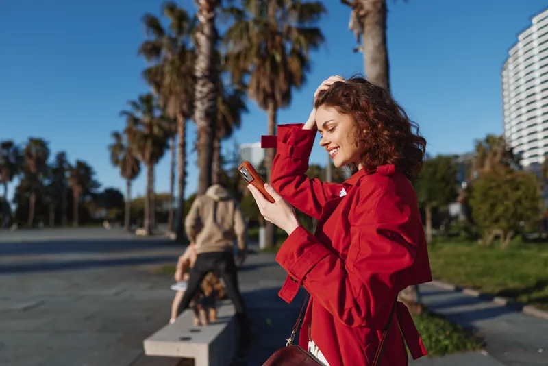 Cheerful woman outdoors in a red coat, smiling at her phone, sunny urban scene with palm trees, stylish moment on a bustling street during golden hour, vibrant surroundings