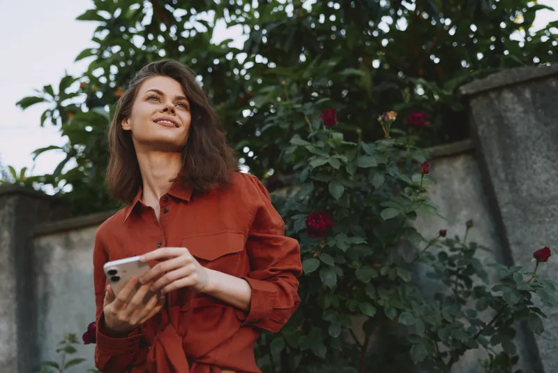 Young woman with shoulder length hair wearing a red shirt smiles while holding a smartphone outdoors among blooming roses