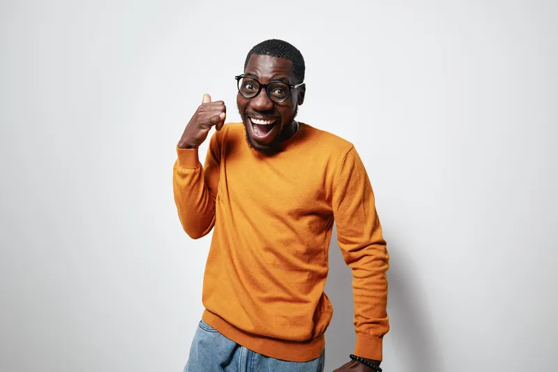 African American man with dark skin wearing glasses and orange sweater smiling and making call me gesture on white background. Happy casual male posing with excitement and joy.