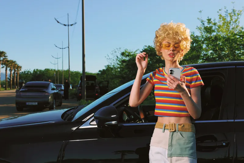 Young woman with curly hair checks her smartphone beside a dark car on a sunny street, wearing a colorful striped top and light skirt for a retro casual lifestyle.