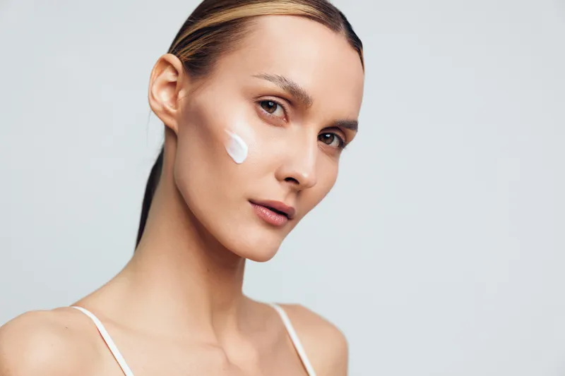 Young woman with clear skin applying face cream, skincare routine, natural beauty, soft lighting, closeup portrait, neutral background, healthy complexion, calm expression