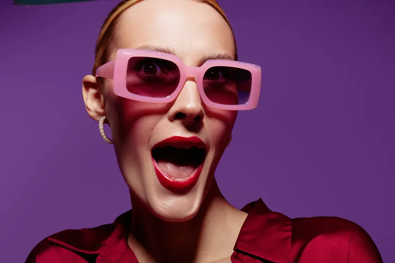 Stylish woman with pink sunglasses and red lipstick expresses surprise on purple background. Fashion portrait with bold makeup and trendy accessories. Concept of beauty and modern style.