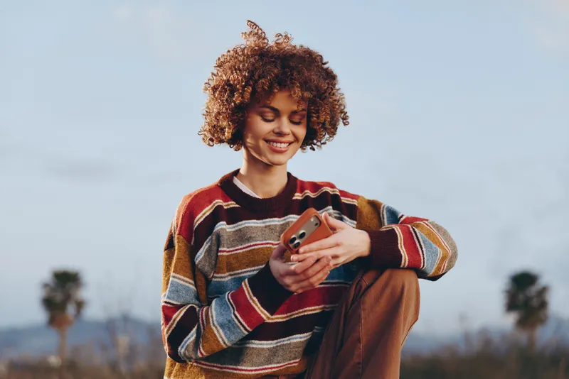 Woman lifestyle outdoors, smiling with curly hair, holding a smartphone, casual striped sweater, relaxed pose, bright sky, soft wind, natural colors, open field background, now