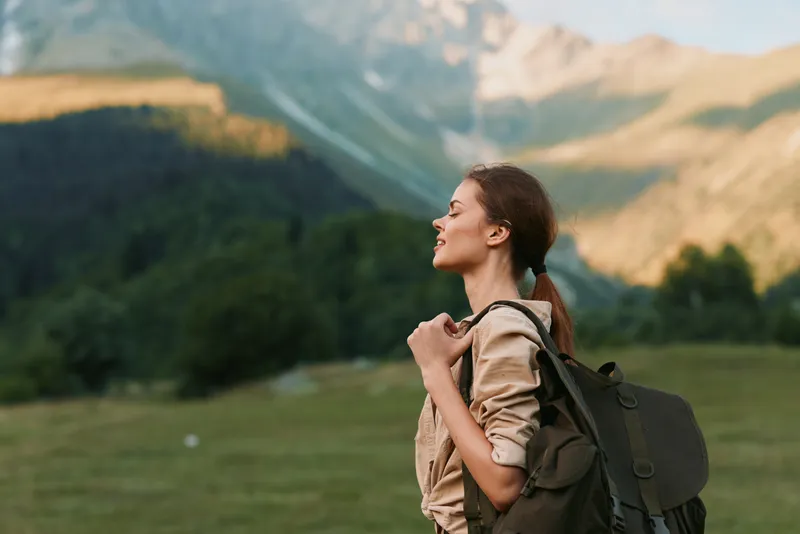 Cheerful woman outdoors, hiking through a sunlit meadow with distant mountains, wearing a backpack, relaxed smile, natural colors, lifestyle moment, calm breeze