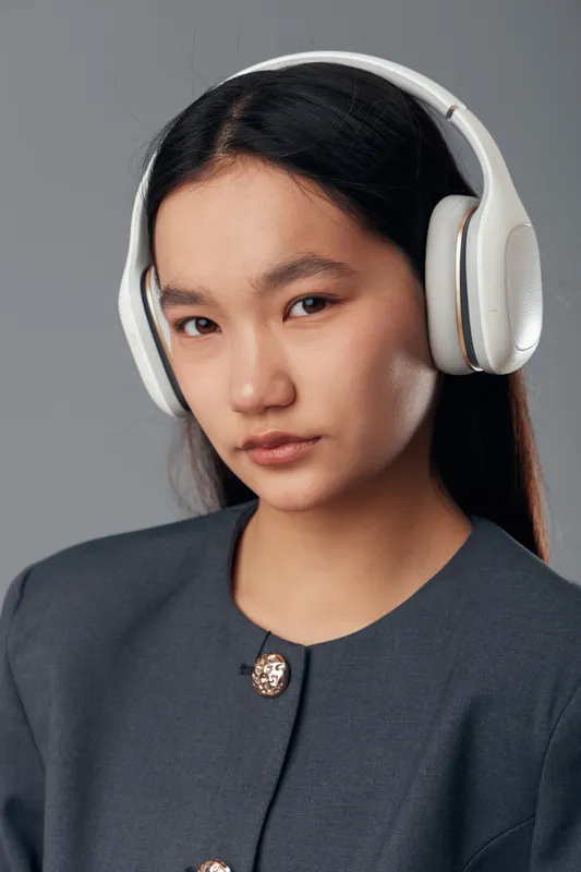 Young woman with long dark hair wearing white wireless headphones and a dark tailored jacket posing with neutral expression on gray background for technology and lifestyle.