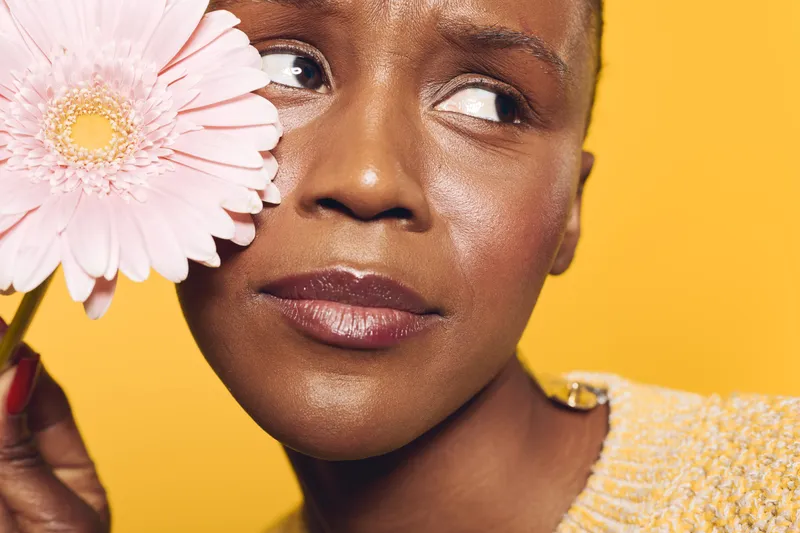 Close-up of a dark skinned African woman holding a pink gerbera flower near her face. The yellow background and soft lighting highlight her natural beauty and calm expression.