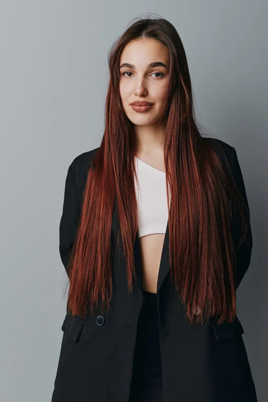 Young woman with long braided hair wearing black blazer and white top standing against gray background. Portrait of confident female with natural makeup and casual pose indoors.